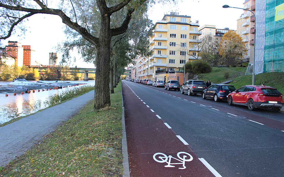 Cykelväg, bilväg och en gångväg av grus med träd och gräs runt om sig. Gångvägen ligger vid vattnet. Parkerade bilar och hus. Foto.
